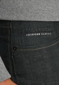 Tamno plave traperice sa zlatnim šavovima, s prednjim džepom i utisnutom oznakom koja glasi "PME Legend American Classic."