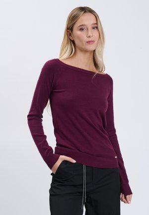 Giovane donna con lunghi capelli biondi che indossa un maglione aderente color bordeaux con spalle scoperte e pantaloni neri, in piedi con una mano in tasca.