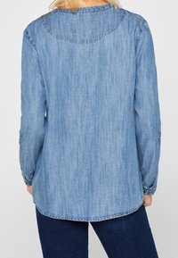 Jasnoniebieska koszula z denimu z długimi rękawami, krzywym hemem i guzikami przy mankietach. Materiał ma miękkie, teksturowane wykończenie.