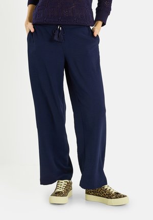 Persona con pantalones anchos azul marino con cintura de cordón y zapatillas deportivas de encaje con estampado de leopardo sobre un fondo blanco liso.