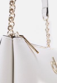 Borsa a mano bianca in pelle con hardware dorato, dotata di chiusura a zip e tracolla a catena. I dettagli notevoli includono un accento con logo dorato.