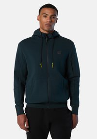 Felpa con zip di un verde scuro, dotata di cappuccio con coulisse, tasche frontali e logo sul petto. Realizzata in tessuto morbido con polsini a coste.