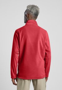 Rode fleece jas met een hoge kraag en lange mouwen. Glad oppervlak, vlakke naden en zijopeningen voor bewegingsvrijheid. Label zichtbaar aan de zoom.