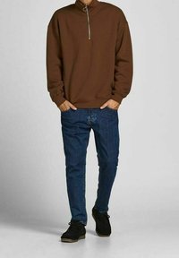 Sudadera marrón con cremallera en el cuello y mangas largas, combinada con jeans azules. Zapatos negros con acabado texturizado y suelas de goma ligeras.