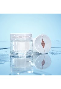 Der Behälter der Magic Water Cream von Charlotte ist aus klarem Glas mit einem weißen Plastedeckel. Er trägt ein kupferfarbenes Logo und Wassertropfen auf einer blauen reflektierenden Oberfläche.