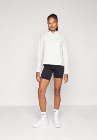 Lagani bijeli jakna s kapuljačom, dugim rukavima i Nike logotipom. Uparena s crnim biciklističkim šortsem. Model nosi bijele tenisice i čarape.