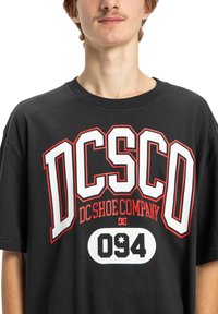 Čierne bavlnené tričko s veľkým grafickým logom v bielej a červenej farbe, s tučnými písmenami "DC" a "SCO" a číslom "094" vo vnútri bielej oválnej plochy.