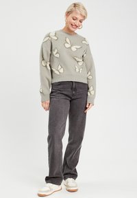 Pull vert clair avec un motif de papillon crème, décolleté rond, épaules tombantes, associé à un pantalon gris foncé et des baskets claires.