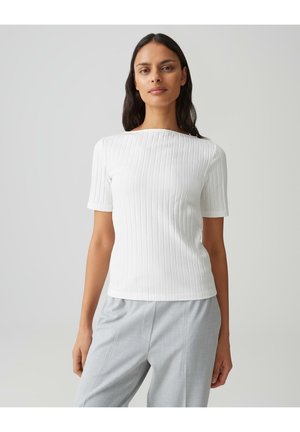 Femme aux longs cheveux bruns portant un haut blanc à manches courtes côtelé et un pantalon gris clair sur un fond uni clair.