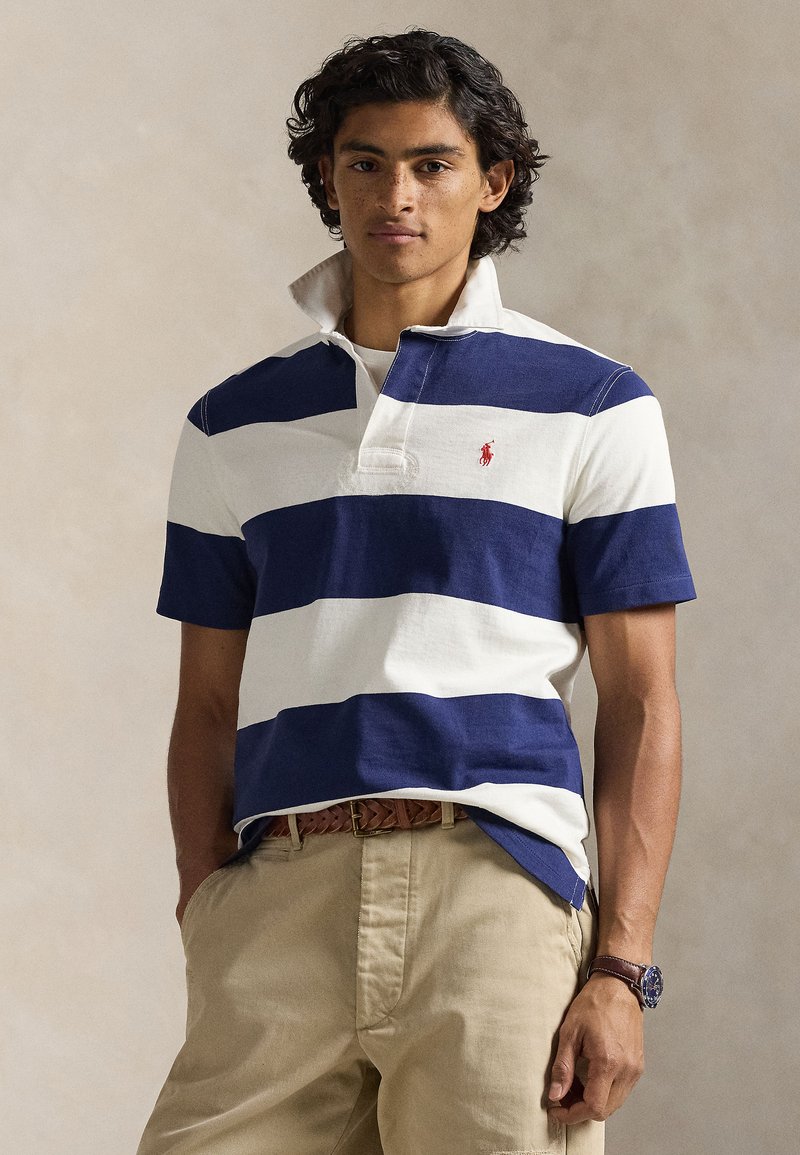 Polo rayé bleu marine et blanc, manches courtes, avec col et un petit logo rouge. Porté avec un pantalon beige et une ceinture tressée.