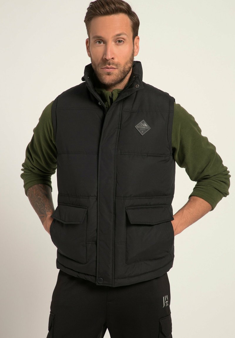 Schwarze Steppweste mit hohem Kragen, zwei Fronttaschen und einem Logo-Patch. Über einem grünen Langarmshirt getragen. Strukturiertes Gewebe.