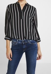Schwarz-weiße gestreifte Bluse mit V-Ausschnitt und hochgekrempelten Ärmeln, kombiniert mit dunkelblauen Jeans.