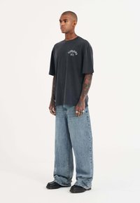 Mørkegrå oversized T-shirt med grafisk print foran, parret med bredbenede lyseblå denimjeans og sorte sko med en tyk sål.
