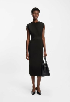 Czarna obcisła sukienka midi z krótkimi rękawami typu "cap" i przemyślanym krojem, zestawiona z czarną torebką i czarnymi szpilkami w szpic.