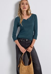 Haut à manches longues teal avec un col rond et une patte de boutonnage à trois boutons, associé à un jean large gris foncé et un tote bag en daim beige.