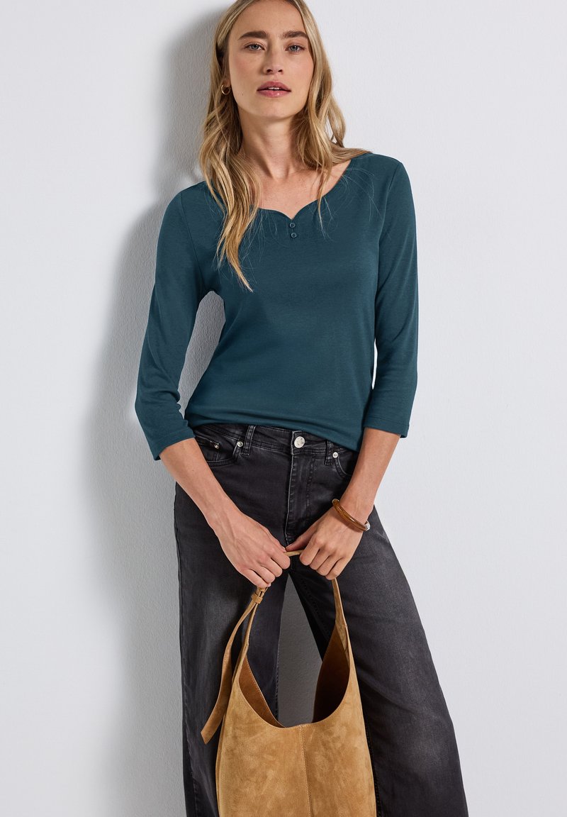 Haut à manches longues teal avec un col rond et une patte de boutonnage à trois boutons, associé à un jean large gris foncé et un tote bag en daim beige.