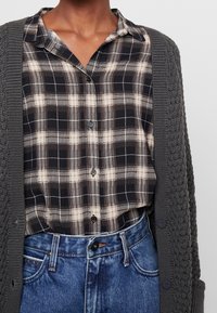 Cardigan grigio scuro a maglia sopra una camicia a bottoni a quadri nera e beige. Abbinato a jeans in denim blu a vita alta.