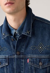 Donkerblauwe spijkerjas met metalen studs op de schouders en voorkant, een klassieke kraag, en twee voorkanten met knoopsluitingen.