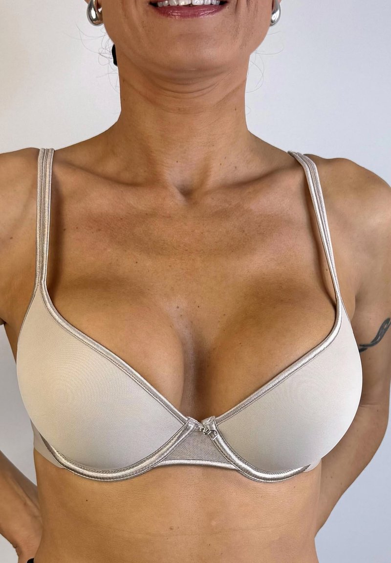 Sujetador beige con tela suave, con un borde de encaje y un diseño con un nudo en el frente. Cuenta con tirantes ajustables y un pequeño cierre central.