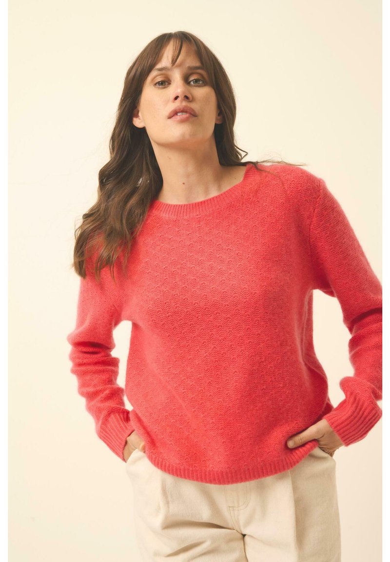 Strickpullover in leuchtendem Koralle, mit Rundhalsausschnitt, langen Ärmeln und einem strukturierten Muster. Kombiniert mit hellen, locker sitzenden Hosen.