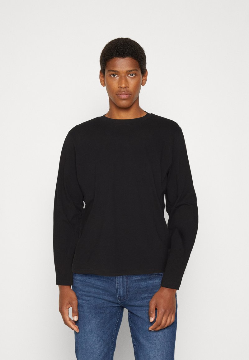 Camiseta negra de manga larga hecha de suave tela de algodón con cuello redondo. Combinada con jeans azules, exhibida sobre un fondo neutro.