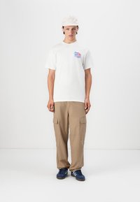 White t-shirt with blue and red graphic, beige cargo pants with side pockets, navy sneakers, and a light-colored cap.