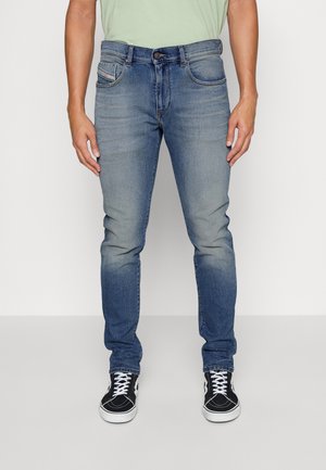 Slim-fit blue denim jeans with a faded wash, five pockets, and a front button closure, paired with black and white sneakers.