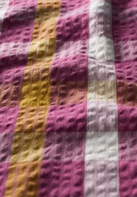 Textile featuring a textured plaid pattern with pink, white, and mustard yellow stripes, highlighted by a slight sheen and varied weave.