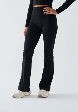 Czarne legginsy z wysokim stanem o kroju dzwonu, wykonane z elastycznego materiału, o gładkiej fakturze i dopasowanym fasonie, zestawione z jasnymi sneakersami.