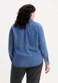 Camicia in denim azzurra con vestibilità rilassata, maniche lunghe e polsini con bottoni. Presenta un orlo arrotondato e un dettaglio sul giromanica posteriore.