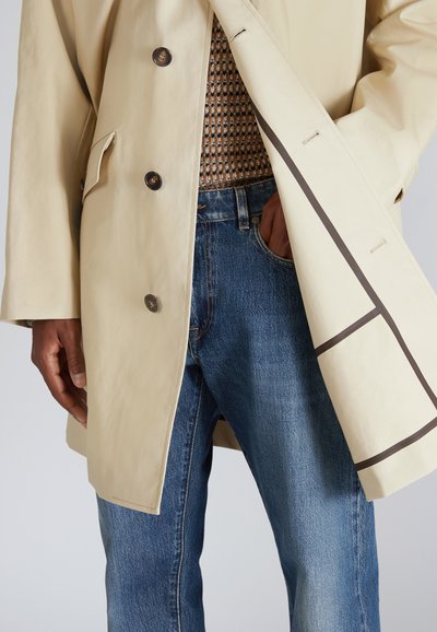 Manteau imperméable beige avec de grands boutons marron, poche latérale et rayures marron, associé à un jean en denim bleu moyen.