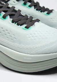 Gros plan sur des chaussures de course gris clair avec des lacets noirs, des oeillets vert menthe, une tige en mesh respirant et une semelle épaisse et coussinée portant l'inscription Hypertech.