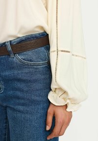 Krämfärgad blus med samlade ärmar, med subtila utskärningar, parat med mörkblå jeans och ett brunt läderbälte.