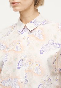 Γυναίκα με πουκάμισο με γιακά και κουμπιά, με παστέλ μοτίβο πεταλούδας και πορτοκαλί τελείες σε ανοιχτό ύφασμα.