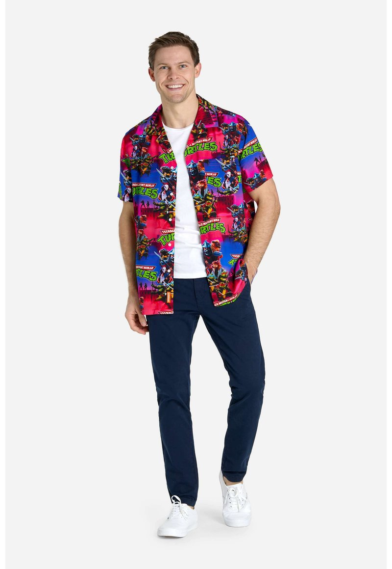 Camicetta a maniche corte con bottoni che presenta un motivo multicolore delle Tartarughe Ninja su uno sfondo rosa, abbinata a pantaloni blu e scarpe da ginnastica bianche.