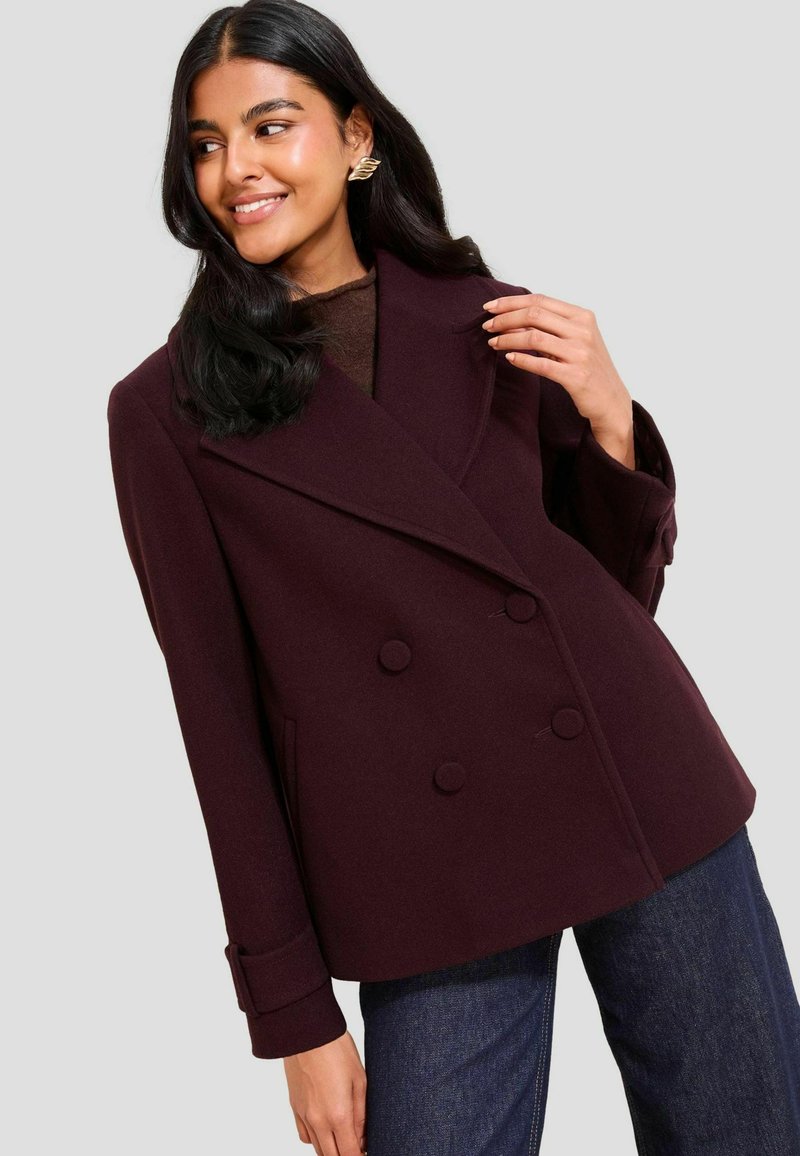 Donna con lunghi capelli scuri che indossa orecchini a onda dorati, un cappotto doppio petto color prugna scuro e jeans blu, sorridendo e guardando a sinistra.