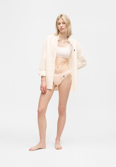 Γυναίκα που φοράει μπεζ ανοιχτό πουκάμισο, αθλητικό σουτιέν και εσώρουχο στο ίδιο χρώμα, όντας ξυπόλητη μπροστά σε ένα απλό λευκό φόντο.