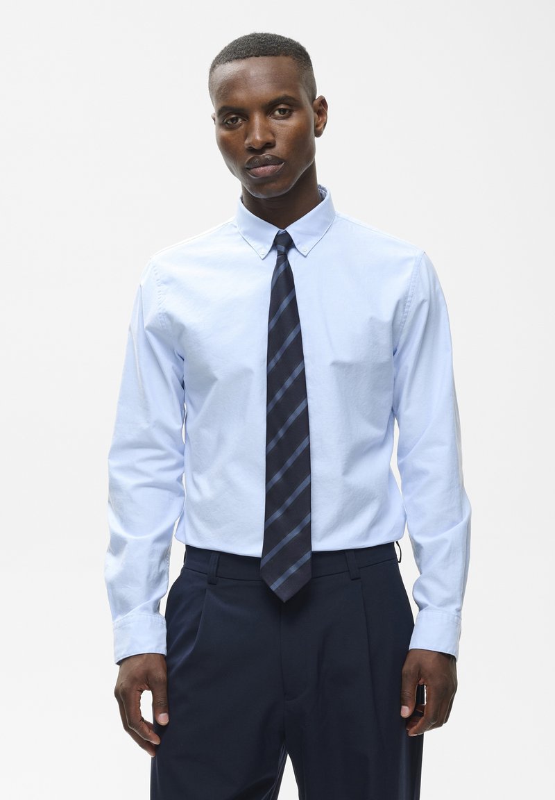 Homme portant une chemise bleu clair à col boutonné et une cravate sombre à rayures diagonales, associée à un pantalon foncé, debout face à l'avant.