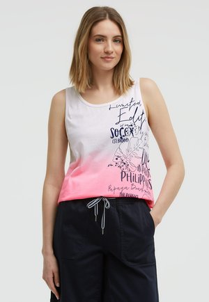 Young woman wearing a white and pink sleeveless top with black text and dark navy drawstring pants standing against a plain background.