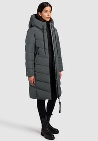 Femme portant un long manteau d'hiver gris foncé rembourré avec une grande capuche, des leggings noirs et des bottines en cuir noires, se tenant devant un fond uni.