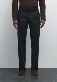 Pantalon noir à coupe droite, confectionné en tissu lisse, avec une fermeture à bouton et des poches latérales. Associé à des chaussures marron foncé.