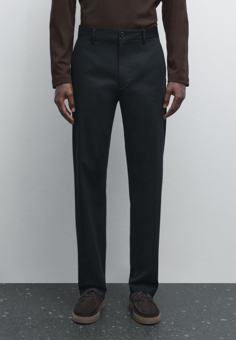 Pantalon noir à coupe droite, confectionné en tissu lisse, avec une fermeture à bouton et des poches latérales. Associé à des chaussures marron foncé.