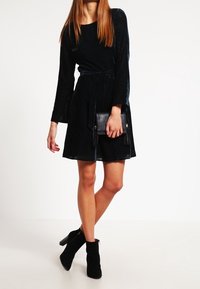Robe noire plissée avec une ceinture à nouer, manches longues et ourlet évasé. Associée à des bottines noires et à un petit sac à main noir.