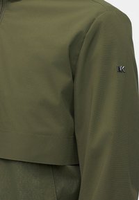 Μανίκι ελιάς πράσινου μπουφάν με μικρό ασημί λογότυπο "MK" στο πάνω μέρος του βραχίονα, που δείχνει την υφή του υφάσματος και τις λεπτομέρειες της ραφής.