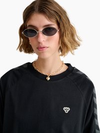 Młoda kobieta o falowanych krótkich włosach, nosząca małe owalne okulary przeciwsłoneczne, złoty naszyjnik z zawieszką w kształcie serca oraz czarną koszulkę z logo pszczoły.