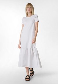 Vestido de algodón blanco con mangas cortas y dobladillo con volantes, que presenta un corte holgado y una textura suave. Combinado con sandalias negras de tiras.