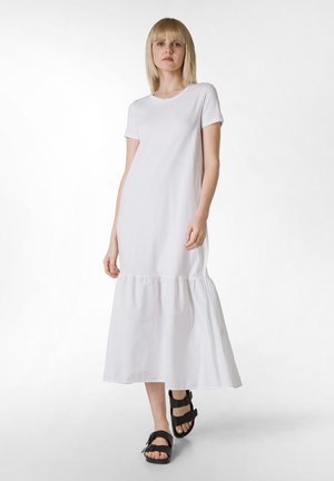 Abito bianco di cotone con maniche corte e orlo a volant, caratterizzato da una vestibilità rilassata e una texture liscia. Indossato con sandali neri con strap.
