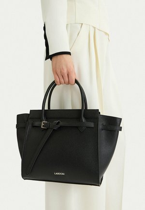 Mano che tiene una borsa in pelle nera con texture, dotata di doppi manici e una cinghia frontale con fibbia, indossata con abbigliamento bianco a pieghe.