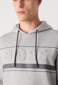 Grey hooded sweatshirt featuring a textured "BOSS" logo and smooth dark grey stripe. Drawstrings are black with plastic tips. Soft fabric texture.