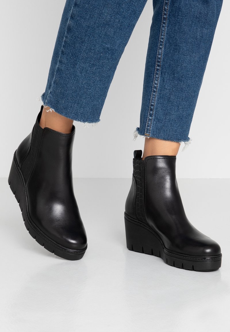 Pieds chaussés de bottines noires à talons compensés associées à un jean bleu court aux ourlets effilochés sur un fond clair uni.